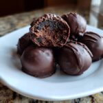 A pile of rich, chocolate-coated Oreo balls on a white plate, with one cut in half showing the dark, dense interior.