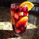 A tall glass filled with vibrant red Christmas sangria, loaded with cranberries, orange slices, and apple chunks.