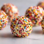 A close-up of one white chocolate-coated sugar cookie truffle covered in bright, multi-colored sprinkles.