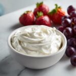 A white bowl filled with creamy fruit dip, surrounded by fresh strawberries and red grapes.