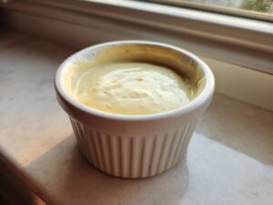 A close-up of creamy garlic sauce served in a small, white, fluted ramekin, sitting on a windowsill.