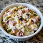 Close-up of creamy hoagie dip loaded with ham, peppers, onions, and olives in a white bowl.