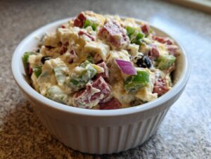 Close-up of creamy hoagie dip loaded with salami, green peppers, and red onion in a white bowl.