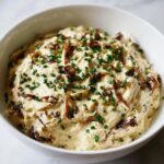 A close-up of a creamy, light brown onion dip topped generously with caramelized onions and fresh green chives.