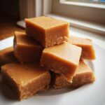 A close-up stack of perfectly cut, creamy penuche fudge squares resting on a white plate near a window.