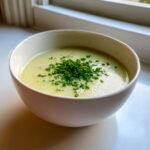 A creamy bowl of potato leek soup topped generously with fresh, chopped chives, sitting near a window.
