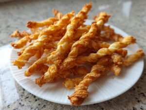 A close-up of a pile of golden brown, crispy cheese straws served on a white ridged plate.