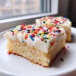Two squares of thick, soft sugar cookie bars topped with white frosting and colorful rainbow sprinkles.