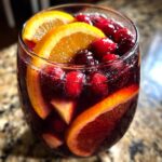 Close-up of a glass filled with dark red holiday sangria, garnished heavily with fresh cranberries and bright orange slices.
