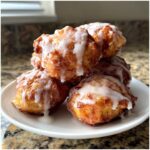 A close-up of several golden brown baked apple fritters stacked on a white plate, drizzled generously with white glaze.