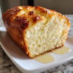 A close-up of a freshly baked beer bread loaf with a shiny, golden-brown crust, resting on a white plate with honey drizzled underneath.