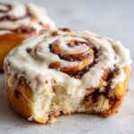 A close-up of a soft, freshly baked cinnamon bun with a bite taken out, covered in thick, white frosting.