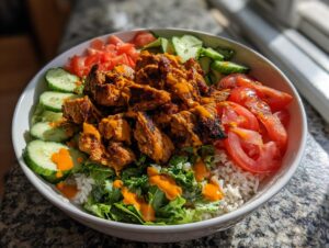 A close-up of a vibrant harissa chicken bowl featuring seasoned chicken pieces over rice, lettuce, cucumbers, and tomatoes, drizzled with orange sauce.