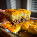A hand holds up a thick slice of John Wayne Casserole showing layers of seasoned ground beef and a fluffy topping covered in melted cheddar cheese.