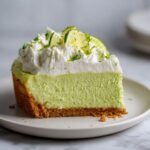 A perfect slice of bright green key lime cheesecake with a graham cracker crust, topped with whipped cream and lime zest.
