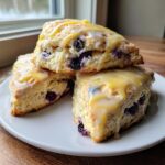 Three freshly baked lemon blueberry scones stacked on a white plate, drizzled with lemon glaze.