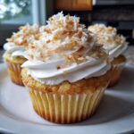 Close-up of moist coconut cupcakes topped with white frosting and toasted coconut flakes.