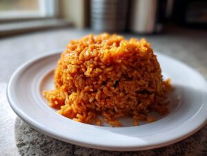 A mound of vibrant, orange-red Nigerian jollof rice served on a white plate, ready to eat.