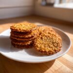 A stack and scattered pieces of golden brown, crispy oatmeal lace cookies resting on a white plate.