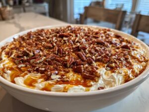 A close-up of a creamy pecan pie dip generously topped with chopped pecans and a swirl of rich caramel sauce.