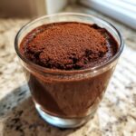 A close-up of rich, dark chocolate mousse served in a small glass, topped generously with cocoa powder.