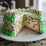 A slice removed from a festive Christmas tree cheesecake showing creamy filling with nuts and green pieces, decorated with green frosting and colorful sprinkles.