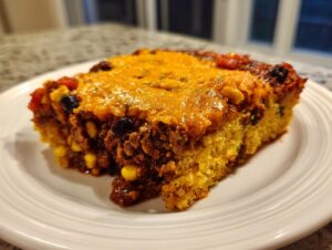 A hearty slice of tamale pie casserole featuring a cornbread base, savory meat filling with corn and beans, topped with melted cheese.