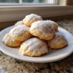 A stack of soft, glazed ricotta cookies topped with coconut flakes on a white plate near a window.