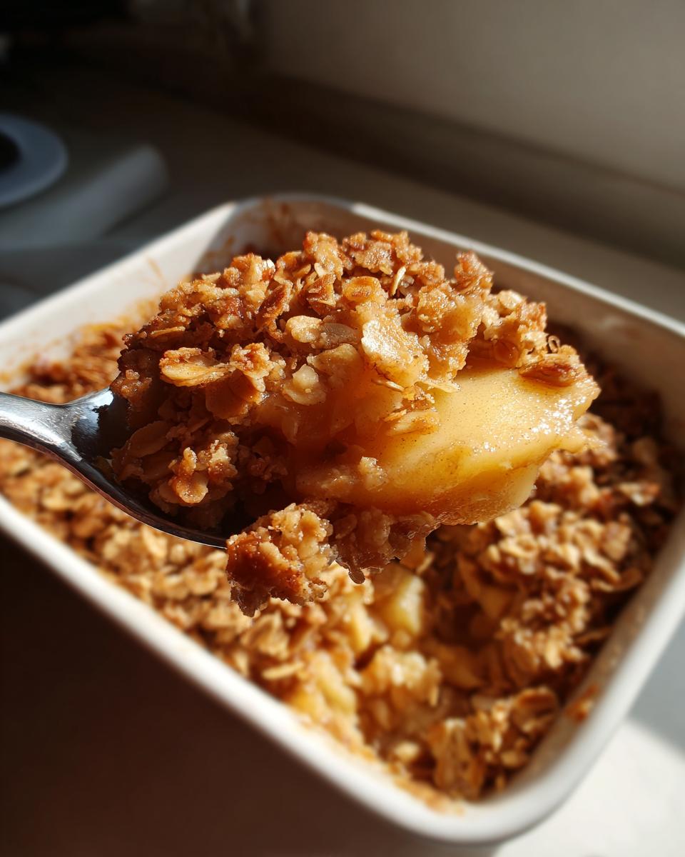 A spoonful of warm apple crumble showing soft cooked apples and crunchy oat topping, lifted from a white baking dish.