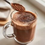 A spoonful of rich brown powder is sprinkled onto a frothy mug of hot cocoa mix.