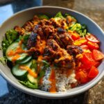 Close-up of a bowl featuring seasoned harissa chicken pieces over rice, topped with sliced cucumbers, tomatoes, and an orange sauce drizzle.