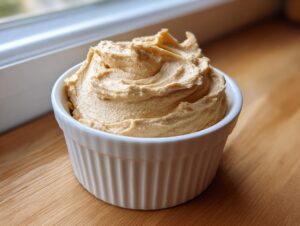 A close-up of freshly whipped honey butter, swirled high in a small white ramekin on a wooden surface.