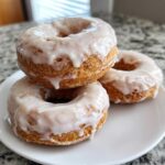 Three freshly made, golden brown cake donuts stacked and drizzled with thick white glaze.