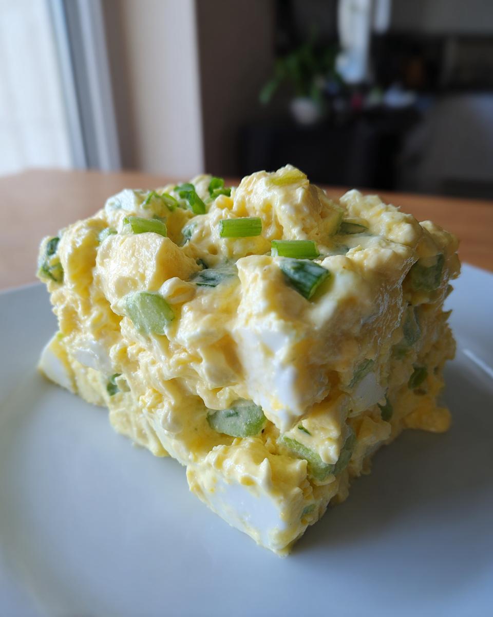 A generous square serving of creamy, chunky egg salad topped with chopped green onions on a white plate.