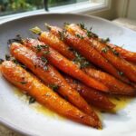 Close-up of shiny, bright orange honey glazed carrots sprinkled with fresh thyme leaves on a white plate.