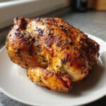 A beautifully roasted whole chicken with golden-brown, herb-crusted skin resting on a white plate.