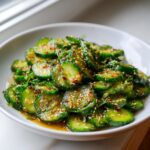 A white bowl filled with glistening, sliced asian cucumber salad tossed in a savory dressing and topped with sesame seeds.