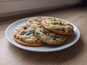 A stack of three chewy, freshly baked m&m cookies resting on a small white plate on a wooden surface.