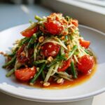 A close-up of a vibrant papaya salad featuring shredded green papaya, cherry tomatoes, green beans, and crushed peanuts.