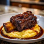 A close-up of a single, tender braised short ribs served over creamy yellow polenta with rich sauce.