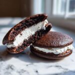 Two rich chocolate whoopie pies, one cut in half showing the thick white cream filling and chocolate layer.