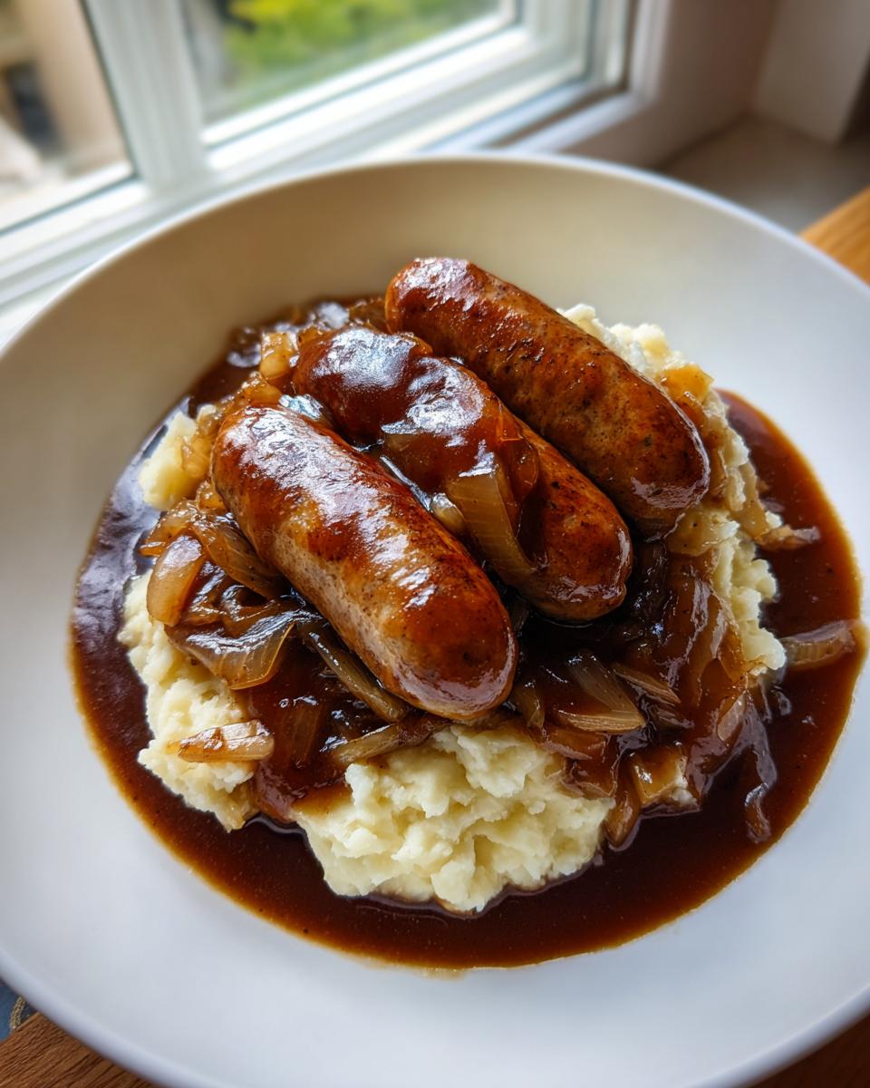 A hearty bowl of bangers and mash featuring three browned sausages over creamy mashed potatoes and smothered in onion gravy.