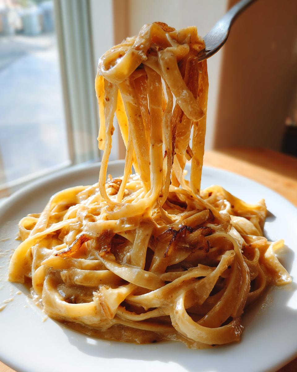 A fork lifts strands of creamy caramelized onion pasta from a white plate, showing rich sauce and browned onions.