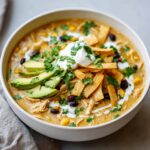 Close-up of a bowl of creamy chicken tortilla soup topped with avocado, sour cream, cilantro, and tortilla strips.