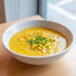 A close-up of a white bowl filled with creamy yellow corn soup, garnished generously with whole corn kernels and fresh chopped chives.