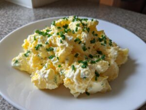 A generous serving of creamy deviled egg potato salad, garnished with fresh green chives, on a white plate.