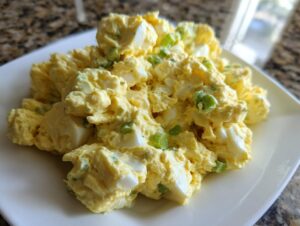 A close-up of creamy, yellow egg salad mixed with chopped celery, served on a white square plate.