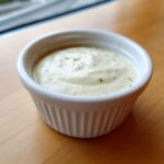 Close-up of creamy homemade garlic aioli recipe served in a small white fluted ramekin on a wooden surface.