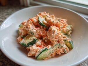 Close-up of a creamy, orange-tinted kani salad mixed with chunks of cucumber, served in a white bowl.