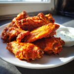 Close-up of crispy, saucy buffalo wings piled on a white plate next to a small bowl of ranch or blue cheese dip.